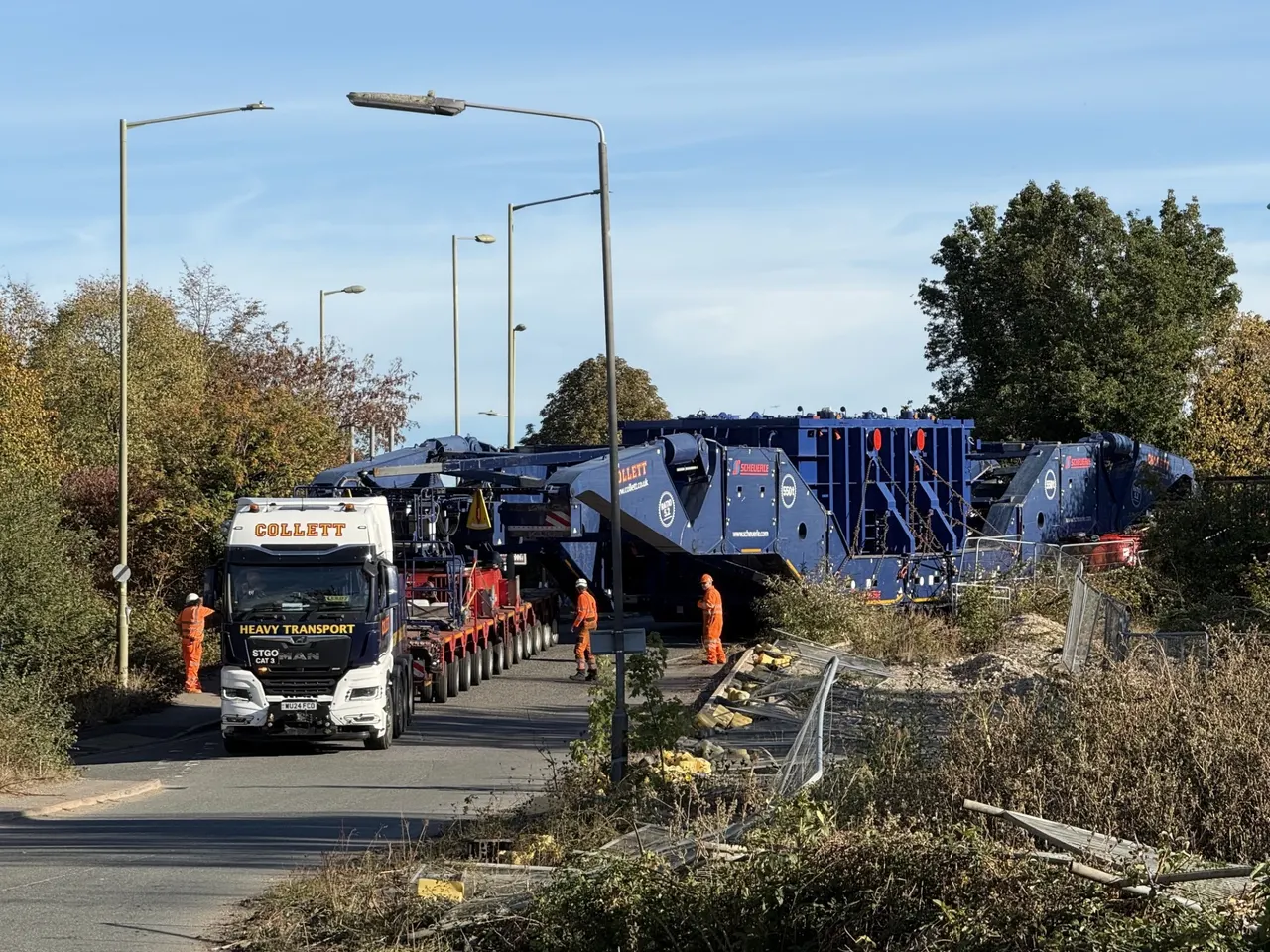 Heavy transport tractor unit leading the girder bridge transporter carrying the transformer, with operatives in orange workwear alongside.