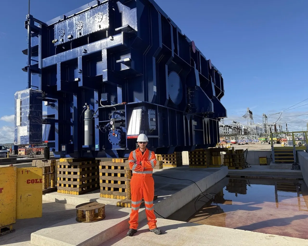 Thomas Featherstone in orange coveralls and a white hard hat in front of the transformer, which rests on timber packing stacks.